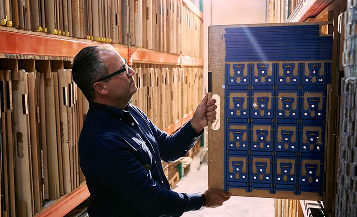 A white man in a dark button-down shirt pulls a board of dies off of a shelf filled with them, holding it up to the sunlight to inspect it.