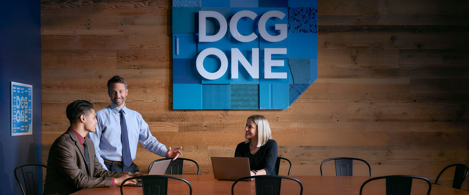 An Asian man, a white man, and a white woman work in a wood-paneled conference room fitted with a big blue DCG ONE sign.