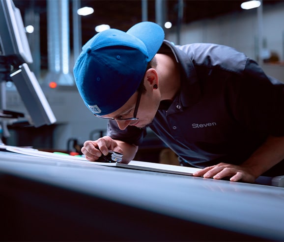 A white man in a blue DCG ONE ballcap and monogrammed polo shirt leans down close to a proof, using a loupe to examine the details.