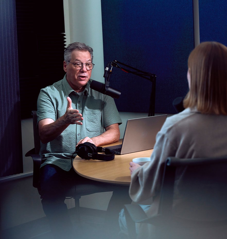A white man with gray hair and long glasses speaks into a microphone to a blonde woman with her back to the camera.