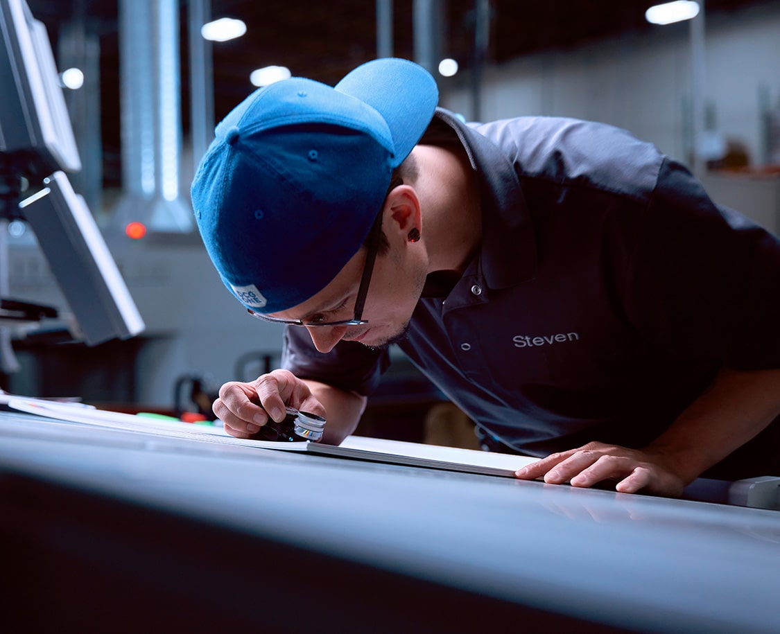 A white man in a blue DCG ONE ballcap and monogrammed polo shirt leans down close to a proof, using a loupe to examine the details.