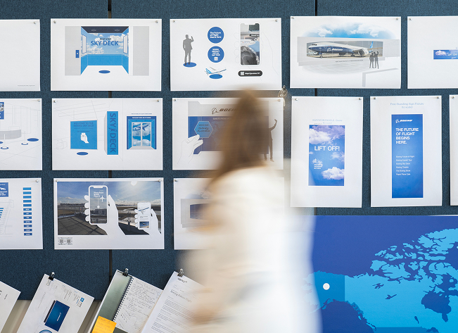 A blonde woman in a long white cardigan is blurred as she moves past a wall covered in tacked-up storyboard screens.